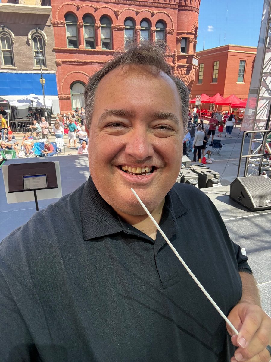 R.J. holding a conductor's baton on an outdoor stage, crowd and historic red-brick building behind him